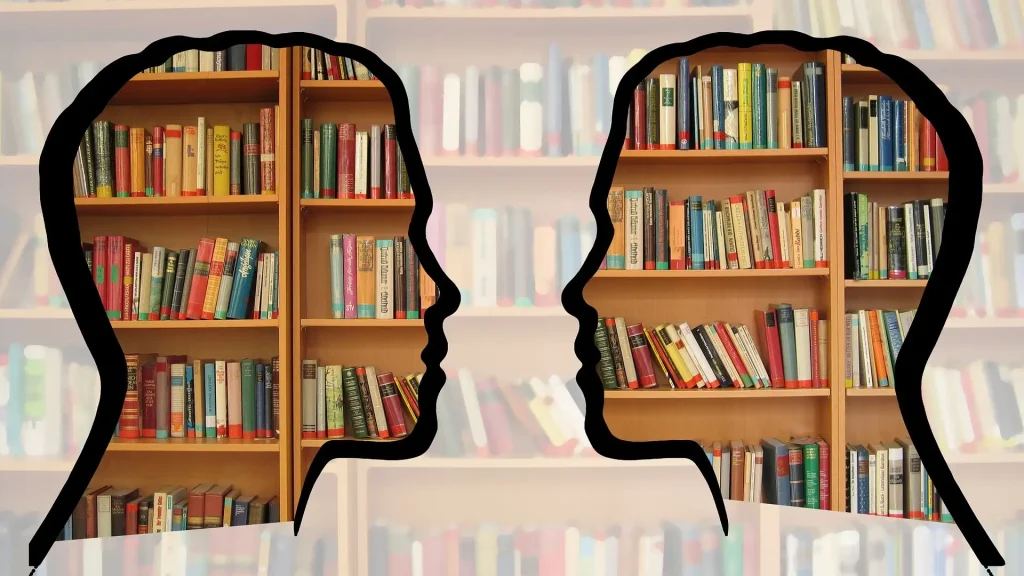 Es ist im Hintergrund ein Bücherregal zu sehen und im Vordergrund zwei Köpfe die sich ansehen.